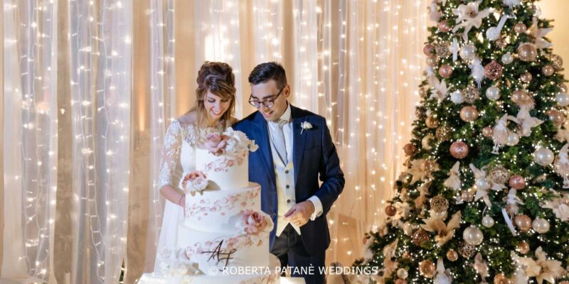 Sposi che tagliano la torta in un matrimonio a tema natalizio con l'albero di Natale e tenda di luci di sfondo. Un matrimonio firmato Roberta Patanè Weddings.